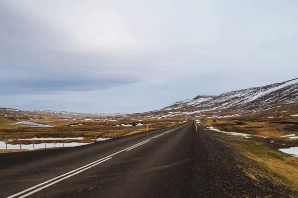 Weg omgeven door heuvels bedekt met sneeuw en groen onder een bewolkte lucht in IJsland — Stockfoto