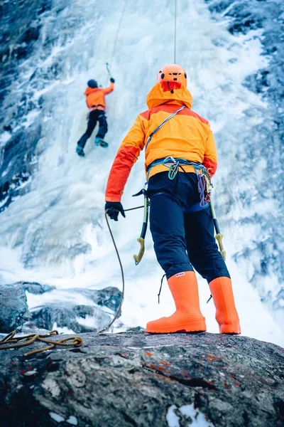 Baixo ângulo de tiro de um alpinista de gelo em pé sobre uma rocha e seu amigo subindo ao fundo — Fotografia de Stock