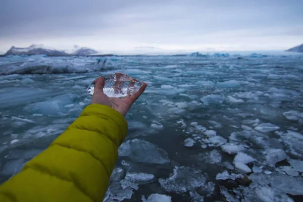 Χέρι κρατώντας πάγο με μια παγωμένη θάλασσα κάτω από ένα συννεφιασμένο ουρανό στην Ισλανδία στο παρασκήνιο — Φωτογραφία Αρχείου