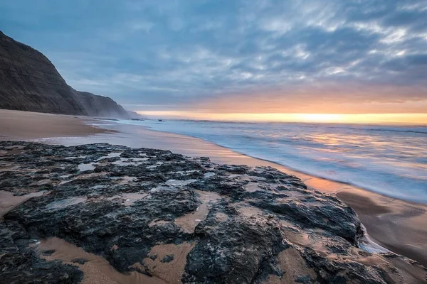 Praia cercada por rochas e o mar sob um céu nublado durante um belo pôr do sol — Fotografia de Stock