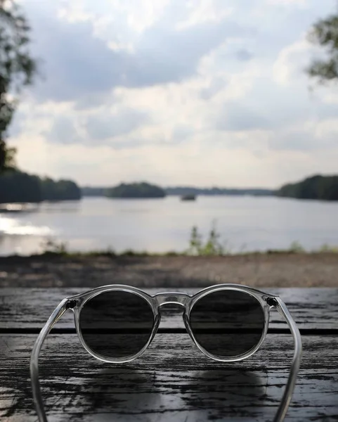 나무 탁자 위에 있는 시원 한 선글라스의 수직 사진 과 뒤에 있는 호수 — 스톡 사진