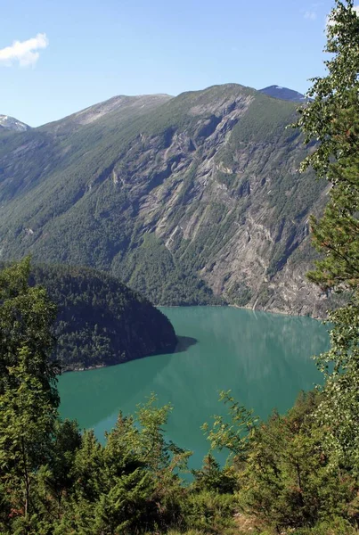 Tiro vertical de um lago no meio de uma montanha florestada abaixo de um céu azul — Fotografia de Stock