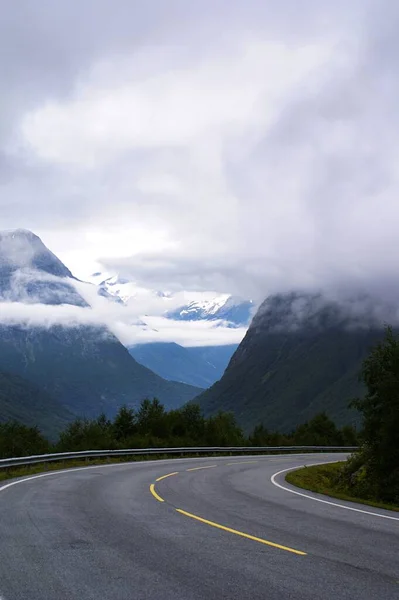 Vertikální záběr na silnici obklopenou vysokými skalnatými horami pokrytými bílými mraky — Stock fotografie