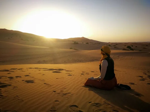 Mujer sentada en el suelo arenoso en el desierto y admirando la puesta de sol — Foto de Stock