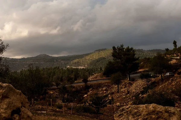 Carretera rodeada de colinas cubiertas de rocas y bosques bajo la luz del sol y un cielo nublado. — Foto de Stock