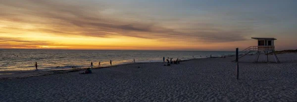 夕日の曇り空の下、人々と海に囲まれたビーチのパノラマ — ストック写真