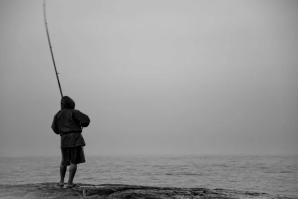 Tir en niveaux de gris d'une personne tenant un poteau de pêche au bord de la mer — Photo