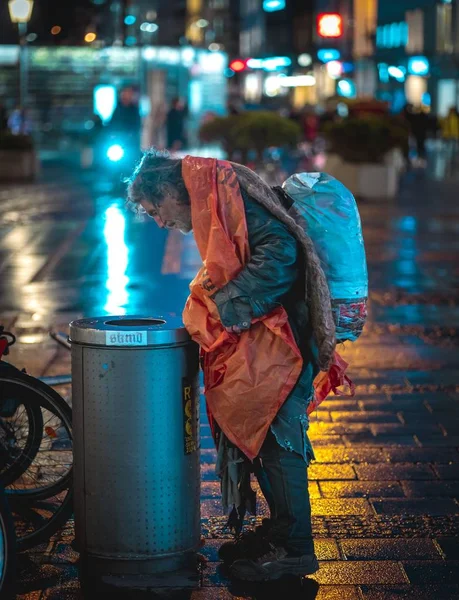 ミュンヘン ドイツ 2020年2月1日 ホームレスの男性がゴミ箱で飲み物や食べ物を探しています ミュンヘンの寒い雨の夜 街の光を反映したぬれた通り — ストック写真