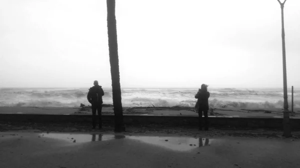 Scala di grigi girato di un uomo e una donna in piedi sulla riva e scattare foto delle onde — Foto Stock