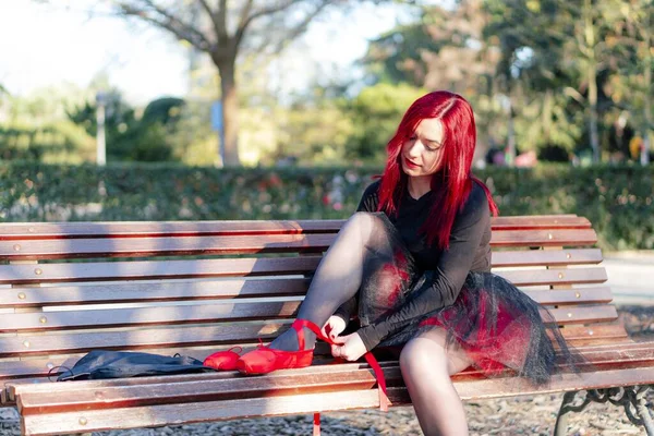 一位漂亮的红头发芭蕾舞演员穿着红点鞋坐在公园的长椅上 — 图库照片