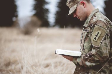 A shallow focus shot of a young soldier reading a bible in a field clipart