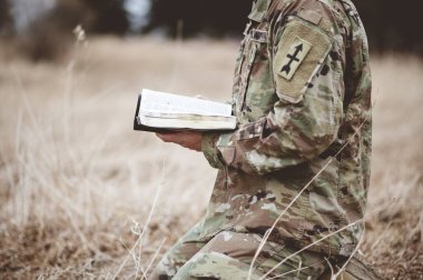 A shallow focus shot of a young soldier kneeling while holding an open bible in a field clipart