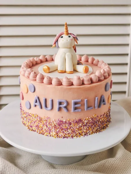A vertical closeup of a birthday cake with a unicorn on the top and the name Aurelia on it on a plate