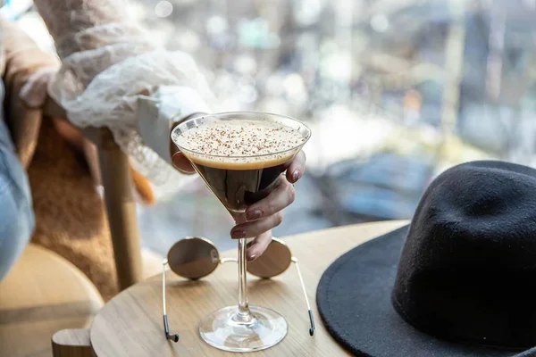 close up view of a woman\'s hand picking a martini glass filled with delicious chocolate coffee martini. sunglasses and black hat on coffee table