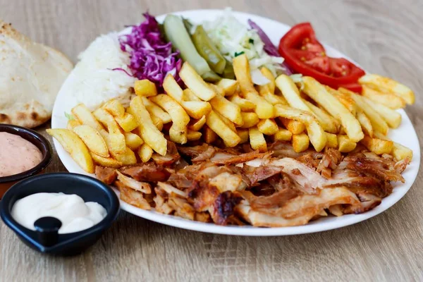 A closeup of Chiken Doner Kebab on a plate with bread, sauce french fries, tomatoes, onion, pickles, cabbage salad on a wooden table