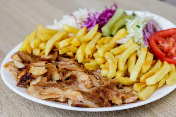A closeup of Chicken Doner Kebab on a plate with french fries, tomatoes, onion, pickles and cabbage on a wooden table