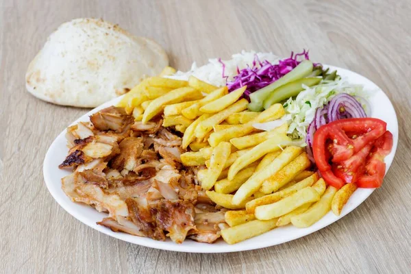 A closeup of Chicken Doner Kebab on a plate with bread, french fries, tomatoes, onion, pickles and cabbage on a wooden table