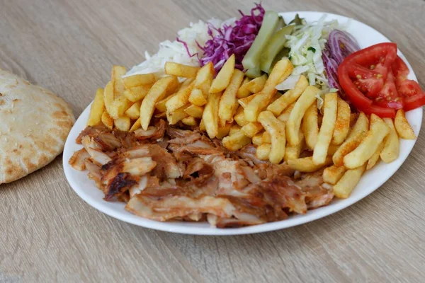 A closeup of Chiken Doner Kebab on a plate with bread, french fries, tomatoes, onion, pickles and cabbage on a wooden table