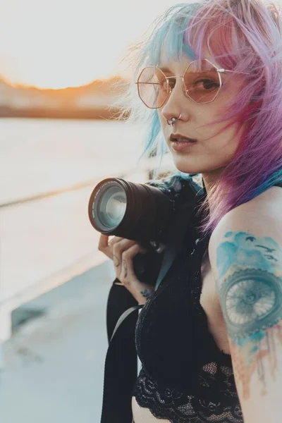 海の近くに立っているカメラを持っているピンクと青の髪と入れ墨を持つ若い女性 — ストック写真