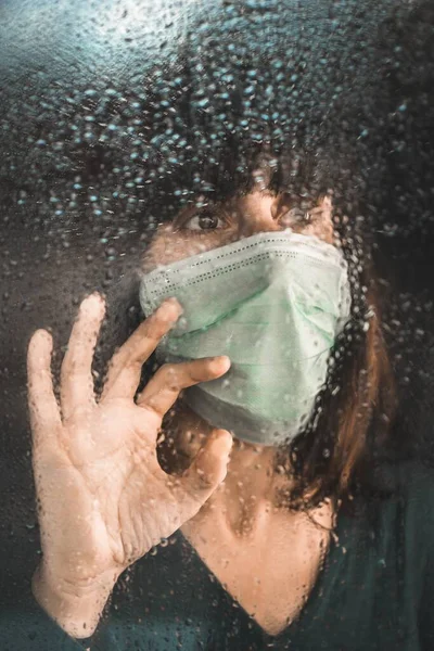 在检疫期间的一个雨天 一个戴着医疗面罩的女人从窗玻璃向外张望 — 图库照片