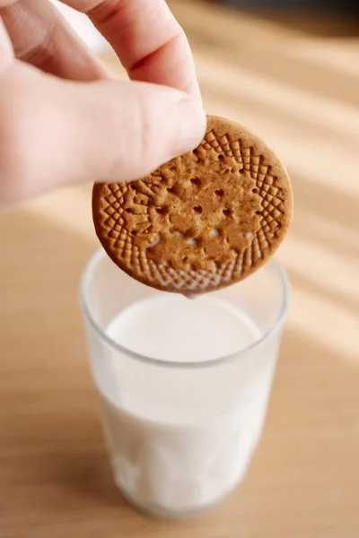 在灯下 一个人在桌上的一杯牛奶里蘸饼干的垂直特写 — 图库照片