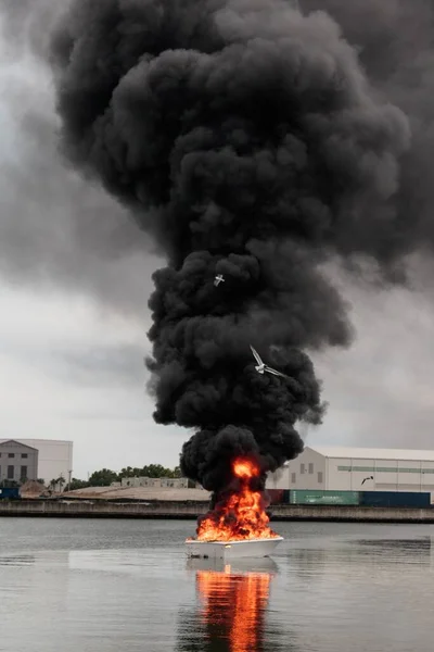 A vertical picture of a burning boat with heavy smoke on the water under a cloudy sky at daytime