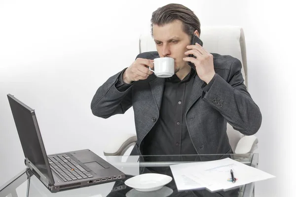 一个年轻人坐在他的办公室里 他在喝咖啡 还想打个电话 他还盯着电脑屏幕看 — 图库照片