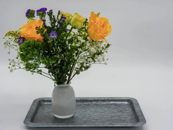A picture of multicolor flowers in a white vase on a silver tray against a grey background