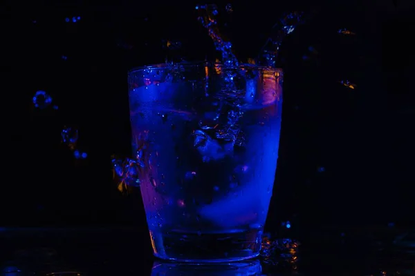 黒い背景のカラフルなライトの下のテーブルの上のガラスに冷たい液体のスプラッシュ — ストック写真