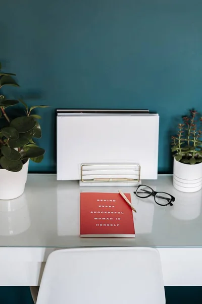 A closeup view of a white desk with white papers, red notebook, plants, pen and glasses in it