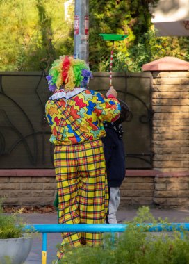 Renkli peruklu ve renkli elbiseli bir palyaço bir çocuğu eğlendiriyor.