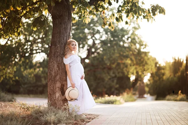 Beautiful blonde pregnant woman 24-26 year old wearing stylish white dress holding straw hat and pregnant tummy outdoors in park. Motherhood. Maternity.