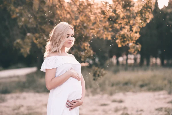 Smiling pregnant woman 20-24 year old wearing white stylish dress standing outdoors over nature background close up. Motherhood. Maternity.