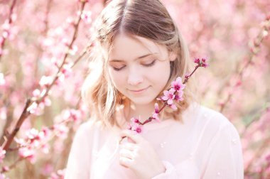 Beautiful smiling blonde girl 16-17 year old posing in peach flowers outdoors. 