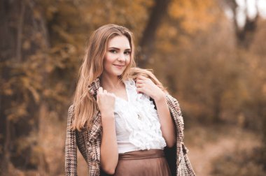 Gülümseyen kadın, 24-26 yaşlarında, uzun sarı saçlı, klasik beyaz bluzlu ve şık ceketli, doğa arka planında poz veren, yakın plan. Kameraya bakıyorum. Sonbahar sezonu. 20 'li. Gençlik.. 