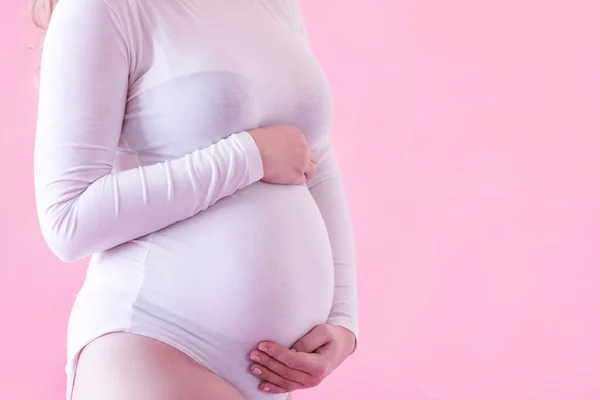 妊娠中の女性25 29歳スタジオクローズアップでピンクの背景の上に手で腹を保持白いボディスーツを着て 健康的なライフスタイルモーターフード産前 — ストック写真