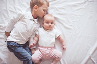 2-3 yaşındaki çocuk, 1 yaşındaki kızı öpüyor. Günlük giysiler giyiyor. Yatakta beyaz çarşafların üzerinde uzanıyor. Kız ve erkek kardeş. Çocukluk. 