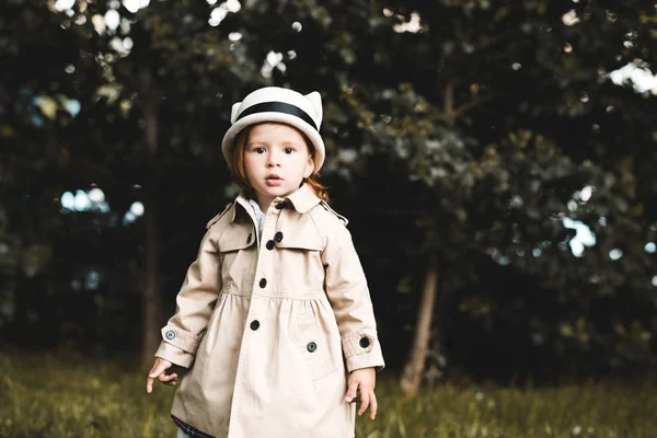 公園の近くを歩くトレンディーなトレンチを身に着けている可愛い女の子 カメラを見て幼少期 — ストック写真