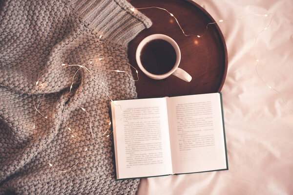 Открытая книга с горячей чашкой черного чая на деревянном подносе с трикотажной одеждой в постели крупным планом. Вид сверху. Пора завтракать. Осенний сезон. Зимние каникулы
. 