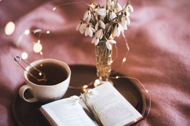 Taze bir fincan siyah çay, açık bir kitap ve yatakta ahşap tepside çiçekler. İyi sabahlar. Bahar mevsimi. 