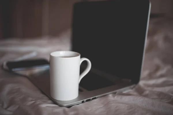 White cup of tea on laptop in bed close up. Quarantine time. Good morning. Brekafast. Working at home.