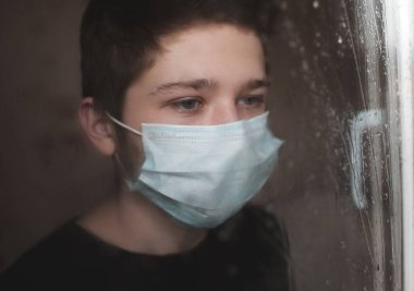 14-16 yaşlarında üzgün bir çocuk. Odasında tıbbi maske takıyor. Yağmurlu pencerelere bakıyor. Üzüntü. Karantina zamanı. Sağlık hizmetleri. Sosyal uzaklık. 
