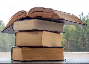 Kalın eski kitaplar pencerenin yanındaki masada yatıyor. Kitaplar birbiri üzerine yatar. En üstteki kitap açık..