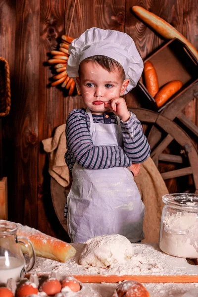 A young child in a chef\'s apron and hat plays the role of a baker.The table is full of clutter. The mood is sublime. There are many ingredients and different things around. Concept for bakery.