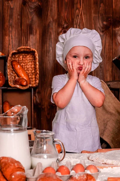 A young child in a chef\'s apron and hat plays the role of a baker.The table is full of clutter. The mood is sublime. There are many ingredients and different things around. Concept for bakery.