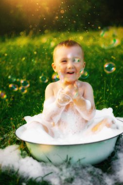 Köydeki mutlu çocuk büyük bir kase köpük ve kabarcığın içinde banyo yapıyor. Mutlu tasasız çocukluk. Yazın kırsal kesimde. Bir sürü köpük ve köpük. Güzel yeşil çimenler. Güneş parlıyor.
