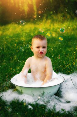 Köydeki mutlu çocuk büyük bir kase köpük ve kabarcığın içinde banyo yapıyor. Mutlu tasasız çocukluk. Yazın kırsal kesimde. Bir sürü köpük ve köpük. Güzel yeşil çimenler. Güneş parlıyor.