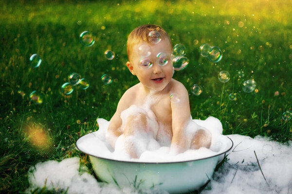 счастливый ребенок в деревне купается в большой миске пены и пузырьков. счастливого беззаботного детства. сельская природа летом. много пены и пузырей. красивая зеленая трава. солнце светит
.