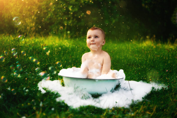 счастливый ребенок в деревне купается в большой миске пены и пузырьков. счастливого беззаботного детства. сельская природа летом. много пены и пузырей. красивая зеленая трава. солнце светит
.