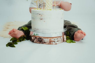 Küçük çocuk beyaz pastanın yanında oturuyor, elleriyle dokunuyor ve tatmaya çalışıyor. Çocuğun pastanın kremasından kirli elleri ve pantolonu var. Hayatın tatlı bir yılı. Tasasız çocukluk.